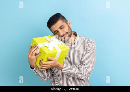 Junger Erwachsener schöner glücklicher Geschäftsmann mit Bart hält umarmt gelb verpackt Geschenkbox, zufrieden, Geschenk zu bekommen, trägt gestreiftes Hemd. Innenaufnahme des Studios isoliert auf blauem Hintergrund. Stockfoto