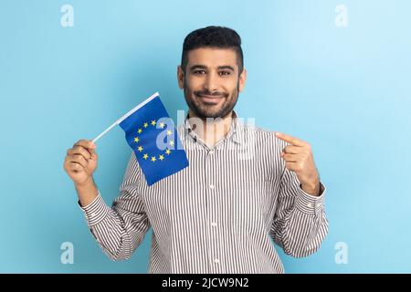 Bärtiger Geschäftsmann, der breit lächelt und die Flagge der Europäischen Union zeigt, Symbol für Europa, EU-Vereinigung und Gemeinschaft, trägt ein gestreiftes Hemd. Innenaufnahme des Studios isoliert auf blauem Hintergrund. Stockfoto