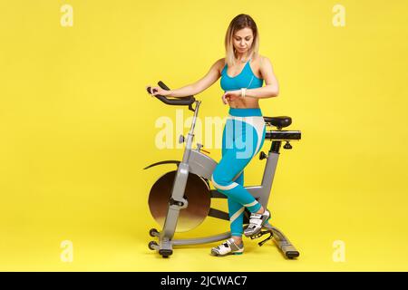 Porträt einer blonden, schönen, athletischen Frau, die in der Nähe des Trainings steht und ihren Fitness-Tracker in blauer Sportswear anschaut. Innenaufnahme des Studios isoliert auf gelbem Hintergrund. Stockfoto