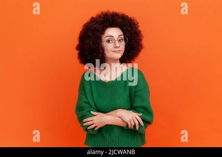 Porträt einer selbstbewussten Frau mit Afro-Frisur, die einen grünen Pullover im lässigen Stil und eine optische Brille trägt, mit gefalteten Armen steht und die Kamera anschaut. Innenaufnahme des Studios isoliert auf orangefarbenem Hintergrund. Stockfoto