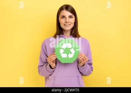 Lächelnde Frau mit dunklem Haar in den Händen halten grünes Recycling singen, Ökologie Konzept, Blick auf die Kamera, trägt lila Hoodie. Innenaufnahme des Studios isoliert auf gelbem Hintergrund. Stockfoto