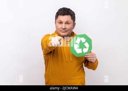 Porträt eines Mannes mit grünem Recycling-Schild, der auf die Kamera zeigt, ruft dazu auf, unseren Planeten vor Umweltverschmutzung zu retten und trägt einen Hoodie im urbanen Stil. Innenaufnahme des Studios isoliert auf weißem Hintergrund. Stockfoto