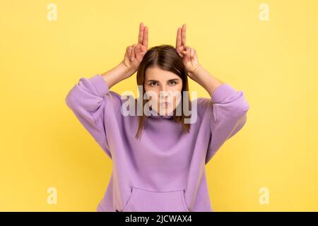 Porträt einer aggressiven Frau, die die Finger über den Kopf hält und Hörner zeigt, arrogant und stur, angriffbereit, mit purpurfarbenem Hoodie. Innenaufnahme des Studios isoliert auf gelbem Hintergrund. Stockfoto