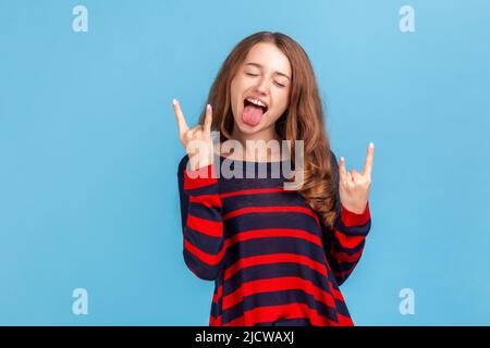 Ja, super. Porträt einer aufgeregten Rockerfrau im Pullover mit Rock and Roll-Handbewegung, verrückten Teufelshörnern mit Fingern und Schreien. Innenaufnahme des Studios isoliert auf blauem Hintergrund. Stockfoto