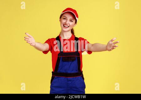 Glücklich lächelnd handliche Frau, die mit ausgestreckten Händen steht und zum Umarmen einlädt, in meine Arme kommt, Overalls und rote Mütze trägt. Innenaufnahme des Studios isoliert auf gelbem Hintergrund. Stockfoto