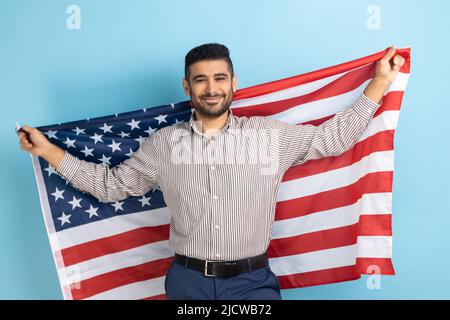 Porträt eines lächelnden Geschäftsmannes, der die US-Flagge hält und mit einem Lächeln auf die Kamera blickt, den Nationalfeiertag feiert und ein gestreiftes Hemd trägt. Innenaufnahme des Studios isoliert auf blauem Hintergrund. Stockfoto
