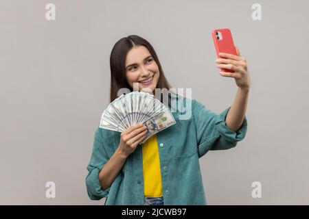 Porträt einer glücklichen reichen Frau, die ein Selfie auf dem Handy mit einem Fan von Dollar-Banknoten in den Händen macht, ihr Gehalt prahlt und eine Jacke im lässigen Stil trägt. Innenaufnahme des Studios isoliert auf grauem Hintergrund. Stockfoto