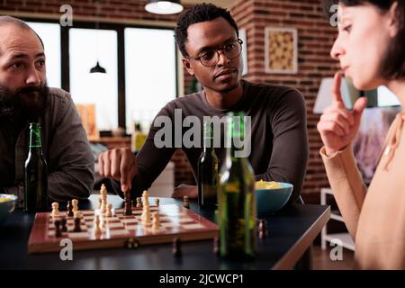 Angespannte multiethnische Menschen sitzen am Tisch, während sie gemeinsam Schach spielen. Multirassische Freunde, die zu Hause im Wohnzimmer sitzen und Strategiespiele und Snacks genießen. Stockfoto