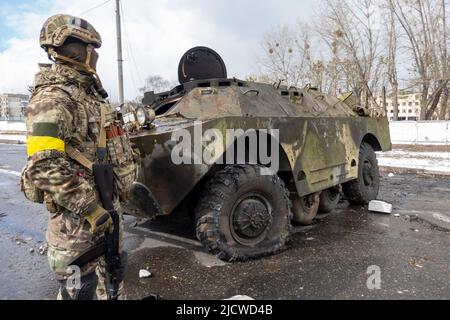 BROVARY, UKRAINE 08. März. Ein bewaffneter ukrainischer Militärangehöriger steht an einem Kontrollpunkt in der Stadt Brovary außerhalb von Kiew neben einem russischen verbrannten gepanzerten Personenträger APC, während die russische Invasion in der Ukraine am 08. März 2022 in Brovary, Ukraine, fortgesetzt wird. Stockfoto