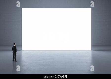 Ein Geschäftsmann, der in einem abstrakten, leeren Galleriensaal mit glänzendem Betonboden auf ein großes weißes blank Poster blickt. Modell Stockfoto
