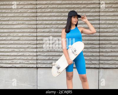 Junge hispanische Frau in blauer Sportbekleidung mit Skateboard, das wegschaut und trendige schwarze Mütze berührt, während sie in der Nähe der rauen Wand des modernen Gebäudes steht Stockfoto