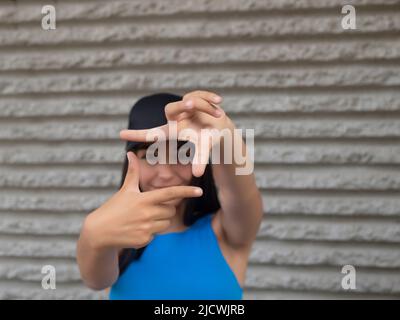 Weicher Fokus einer jungen Frau mit dunklem Haar, die durch den Fingerrahmen auf die Kamera schaut, während sie am Wochenende auf der Straße der Stadt gegen eine raue Wand steht Stockfoto