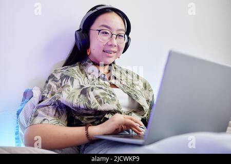 Porträt eines lächelnden Teenagers mit Hosenträger, der in einem gemütlichen Zimmer mit Neonlicht auf dem Bett sitzt Stockfoto