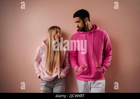 Fashion Studio Portrait eines jungen Biracial Paares im Hoodie, das sich anschaut und auf rosa Hintergrund posiert. Stockfoto