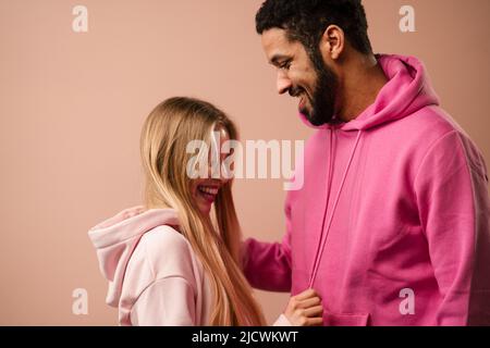 Romantisches Modestudio-Portrait eines jungen Biracial-Paares verliebt in Hoodie posiert auf rosa Hintergrund. Stockfoto