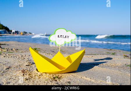 Kleines gelbes Papierboot mit Stick und Sprechblase mit Wort RELAX steht am Sandstrand in der Nähe von Meerwasser und Meereswellen mit weißem Schaum aus der Nähe. Konzept von Reisen, Tourismus, Urlaub, Urlaub Stockfoto