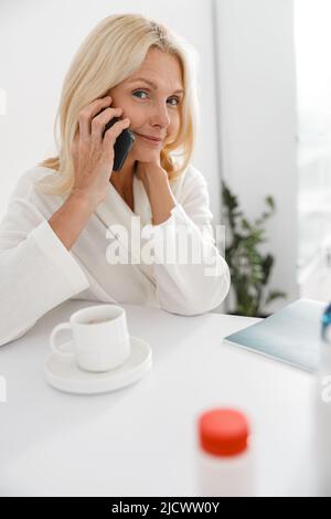 Vertikale Aufnahme einer älteren Dame, die auf dem Smartphone im modernen Kosmetologiezentrum sitzt Stockfoto