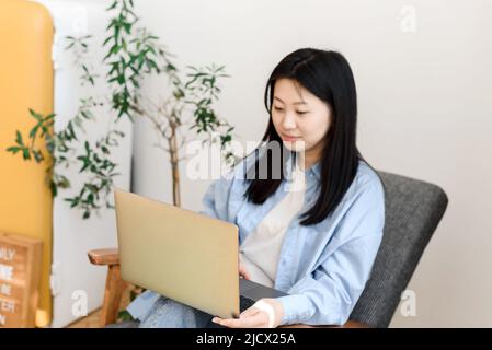 Eine junge asiatische Frau arbeitet hinter einem Laptop und sitzt auf dem Stuhl des Hauses. Koreanische Frau genießt Online-Shopping von zu Hause aus. Stockfoto