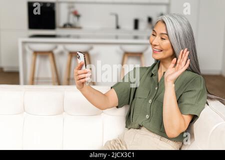 Glücklich lächelnde, reife Frau, die im Wohnzimmer sitzt, sich entspannt und per Videoverbindung mit ihrer Freundin oder Familie spricht, ältere Frau, die Mobiltelefon hält und die Hand winkt Stockfoto