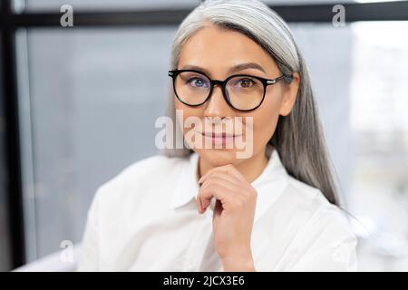 Porträt einer älteren grauhaarigen Ärztin, die am Schreibtisch im Büro sitzt und die Kamera anschaut. Besetzung von Senioren Konzept Stockfoto
