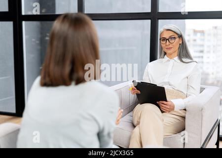 Leitender grauhaariger Therapeut drückt einer verärgerten Patientin während der Therapiesitzung Unterstützung und Empathie aus. Konzept der psychischen Gesundheit Stockfoto