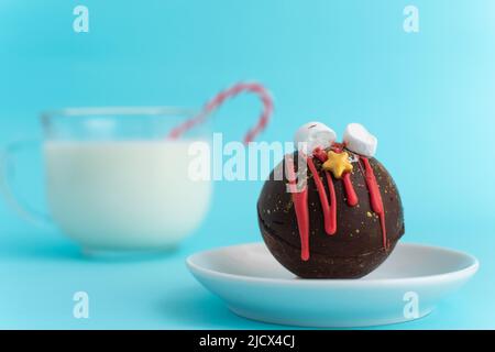Schokoladenbombe mit Marschmalchen und Weihnachtsschmuck auf einem weißen Teller vor einem verschwommenen Bild eines Glases Milch. Blauer Hintergrund. Hochwertige Fotos Stockfoto