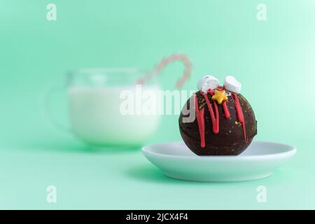 Schokoladenbombe mit Marschmalchen und Weihnachtsschmuck auf einem weißen Teller vor einem verschwommenen Bild eines Glases Milch. Grüner Hintergrund. Hochwertige Fotos Stockfoto