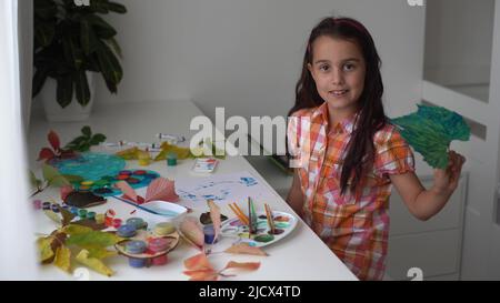 Nahaufnahme eines kleinen Mädchens, das am Tisch sitzt und die Blätter mit Pinsel und Aquarellen zeichnet. Kleines Mädchen, das zu Hause gefallene Blätter malt. Herbst Stockfoto