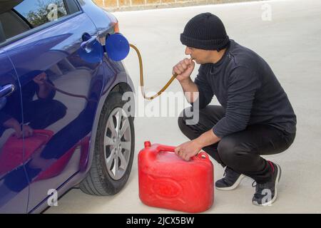 Bild eines Diebes ganz in Schwarz gekleidet, der mit einem Gummischlauch Benzin aus einem geparkten Auto saugt. Verweis auf den aktuell teuren Kraftstoff. Stockfoto