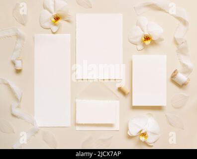 Hochzeitskarten in der Nähe von weißen Orchideenblumen und Seidenbändern auf hellgelber Draufsicht, Mockup. Romantische Szene mit leerer Karte flach legen, Platz für Text. Stockfoto