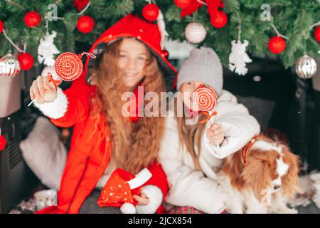 Porträt von zwei Schwestern Mädchen und Hund Kavalier König charles Spaniel Hund mit roten Süßigkeiten Lollipop unter Weihnachtsdekoration im Kofferraum, Kinder haben Stockfoto