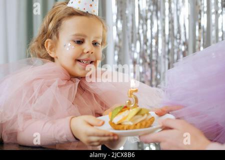 Geburtstag ist fünf Jahre alt. Charmantes, festlich gekleidetes, lächelndes Mädchen nimmt Teller mit Geburtstagskuchen und Nummer fünf auf. Die glücklichen Emotionen der Kinder Stockfoto