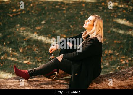 Charmante Frau in warmen, gemütlichen Kleidern geniesstSonnenschein und Wärme setzt ihr Gesicht in die Sonne, die auf einem umgestürzten Baum im Herbstpark sitzt. Herbstporträt. Erholung Stockfoto