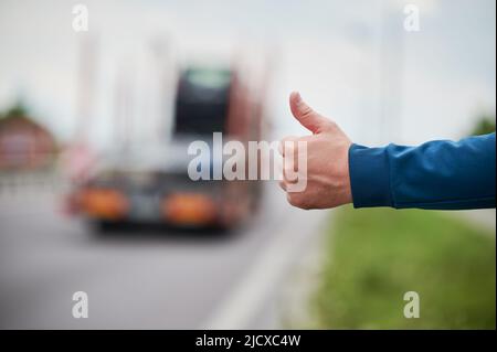 Nahaufnahme der Menschenhand, die am Straßenrand anhält. Männliche Hand zeigt Daumen nach oben Geste im Freien auf unscharfem Hintergrund. Anhalter, Anhalter, Auto-Stopp-Konzept. Stockfoto