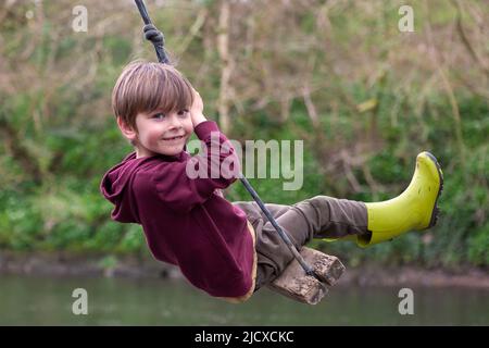 Fünfjähriger Junge, der auf einer Seilschaukel über einem Fluss spielt. MODELL FREIGEGEBEN Stockfoto