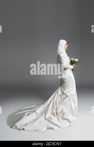 Seitenansicht der glücklichen muslimischen Frau in wunderschönen Hochzeitskleid stehen mit Blumenstrauß auf grau Stockfoto
