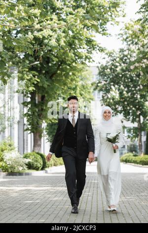 Glücklicher Bräutigam hält die Hände mit lächelnder muslimischer Braut mit Brautstrauß und geht zusammen Stockfoto