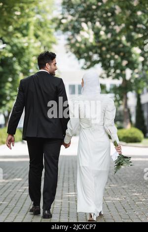 Glücklicher Bräutigam, der die Hände mit einer muslimischen Frau mit einem Hochzeitsstrauß hält und zusammen geht Stockfoto