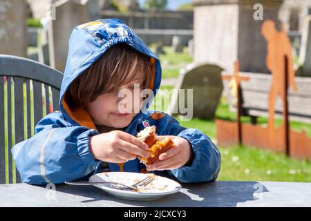 Der kleine Junge, 4 Jahre alt, genießt an einem kühlen Tag in Portchester Castle, Hampshire, Großbritannien, einen Kuchen in einem Café im Freien. MODELL FREIGEGEBEN Stockfoto