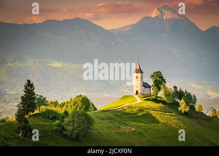 Jamnik, Slowenien - Luftaufnahme der wunderschönen Kirche auf dem Hügel, herrlicher goldener Sonnenuntergang über den Kamnik-Savinja Alpen, Skofja Loka Stockfoto