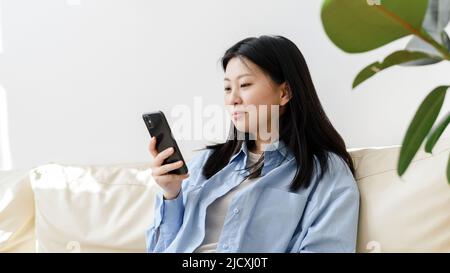 Junge asiatische Frau, die sich auf dem Sofa im Wohnzimmer entspannt und die Nachrichten auf ihrem Smartphone durchstöbert. Koreanische Dame schreibt Nachrichten auf moderne Handy Stockfoto