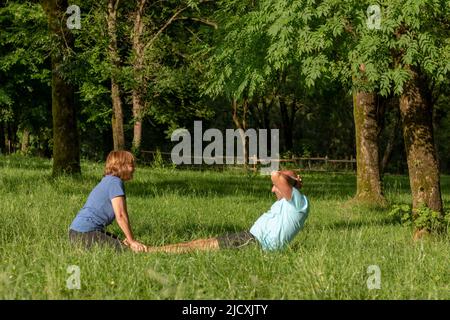 Mann und Frau machen gemeinsam Sit-ups und helfen sich gegenseitig, umgeben von Natur mit an sonnigen Tag Stockfoto