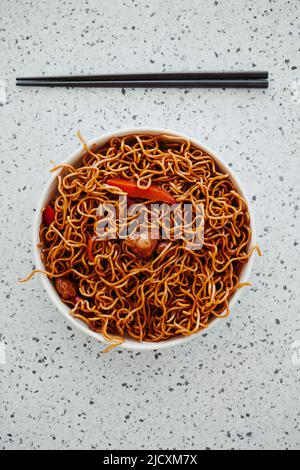 Blick in den hohen Winkel von einem Paar schwarzer Essstäbchen und einem weißen Teller mit einigen Yakisoba-Hühnernudeln auf einer weiß melierten Steinoberfläche Stockfoto