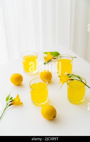 Gläser mit Limonade in der Nähe von frischen Zitronen und gelben Alstroemeria-Blüten auf weißer Tischplatte Stockfoto