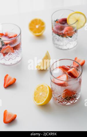 Facettierte Gläser mit Fruchttonikum neben gehackten Erdbeeren und Zitronen auf weißer Tischplatte Stockfoto