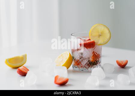 Glas mit kaltem Erdbeertonikum in der Nähe von gehackten Zitronen und Eiswürfeln auf weißer Oberfläche und grauem Hintergrund Stockfoto