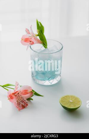Glas mit vereisten Tonic Water in der Nähe von rosa Alstroemeria-Blüten und Kalk auf weißer Oberfläche Stockfoto