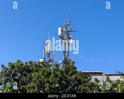 GSM-Antennenturm, Kopierplatz. Telekommunikationstechnologie für 5G, 4G, 3G. Mobiltelefon, TV-Antennen, Dach hinter grünem Baum. Sonniger blauer Himmel Stockfoto