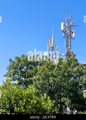 GSM-Antennenturm, Kopierplatz. Telekommunikationstechnologie für 5G, 4G, 3G. Mobiltelefon, TV-Antennen, Dach hinter grünem Baum. Sonniger blauer Himmel Stockfoto