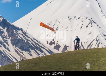 18.05.2022. Gudauri, Georgien. Junger Mann, der an einem sonnigen Sommertag auf einer Bergwiese Fahrrad fährt, schneebedeckte Kaukasus-Berge im Hintergrund. Hochwertige Fotos Stockfoto